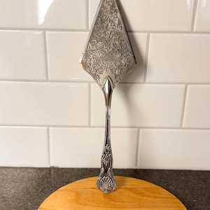 Puede incluir: Una espátula plateada para pasteles con un diseño grabado en la hoja y el mango. La espátula está sobre una tabla de cortar redonda de madera. El fondo es una pared de azulejos blancos.