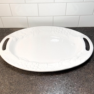 May include: White oval serving platter with an embossed grape and leaf design around the rim. The platter has two handles and is made of ceramic. Suitable for serving appetizers or desserts.