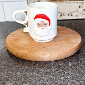 Pode incluir: Uma caneca de cerâmica branca com um rosto 3D do Pai Natal, chapéu vermelho e barba branca. A caneca tem uma pega e decoração de azevinho. Está sobre uma tábua de cortar redonda de madeira.