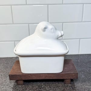 May include: A white ceramic butter dish with a pig-shaped lid, resting on a small, dark wooden tray. The dish has a rectangular base and a rounded top. The background features white subway tiles.