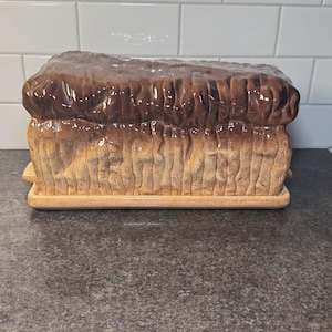 May include: A ceramic bread bin in the shape of a loaf of bread. The top is glazed in a dark brown colour, while the sides and base are a lighter tan. The bin has a rectangular shape with vertical ridges on the sides.