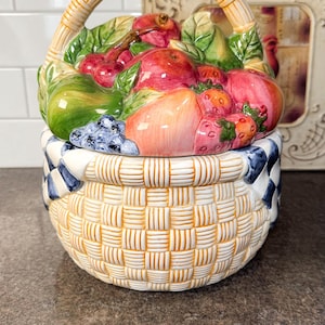 Puede incluir: Un recipiente de cerámica en forma de cesta con tapa, lleno de coloridas frutas esculpidas. La cesta tiene un diseño tejido en amarillo y blanco, con un patrón de cuadros azules y blancos alrededor del borde. El asa también está tejida.