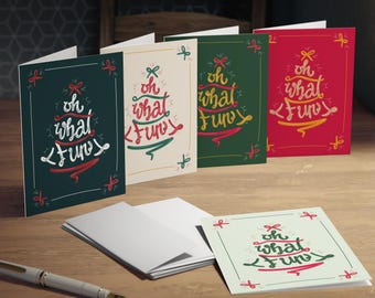 May include: A collection of greeting cards with the phrase "oh what fun" in a festive design. The cards are in various colors, including green, cream, and red, with matching envelopes and a pen on a wooden surface.