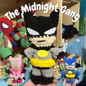 Puede incluir: Muñeco de Batman tejido a ganchillo en gris, negro y amarillo, con detalles blancos en los ojos. El muñeco tiene un cinturón amarillo y un símbolo de murciélago en el pecho. El texto "The Midnight Gang" está en la parte superior. Otros muñecos de superhéroes tejidos a ganchillo están en el fondo.