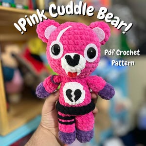 Pode incluir: Um urso de crochê rosa feito à mão com olhos pretos, um focinho branco e um desenho de coração partido preto no peito. O urso tem detalhes roxos nos braços e pernas. O texto na imagem diz "!Pink Cuddle Bear!" e "Pdf Crochet Pattern."