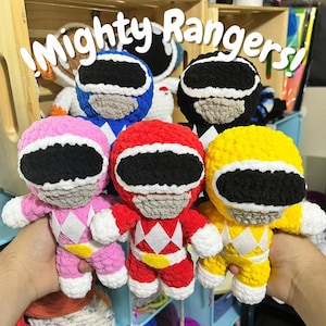 May include: A collection of handmade crochet dolls, each representing a Power Ranger. The dolls are in various colors: pink, red, yellow, blue, and black. The text "Mighty Rangers!" is written in white cursive above the dolls.