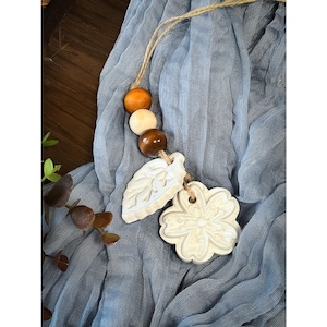 May include: A decorative hanging ornament featuring three round wooden beads in shades of light tan, medium brown, and dark amber, attached to a jute twine string. Two white clay charms, one shaped like a leaf and one like a flower, hang below.
