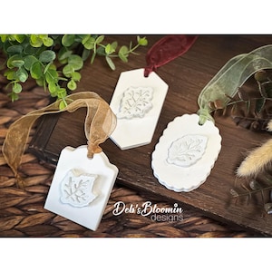 May include: Three white ceramic ornaments with embossed leaf designs, each featuring a different shape: a scalloped oval, a pointed hexagon, and a rounded square. Each ornament is attached to a colored ribbon in gold, red, or green.