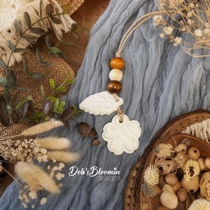May include: A decorative hanging ornament featuring a natural jute cord strung with a smooth orange bead, a light wood bead, and a dark brown bead, followed by a white ceramic leaf charm and a white ceramic flower charm with embossed details.