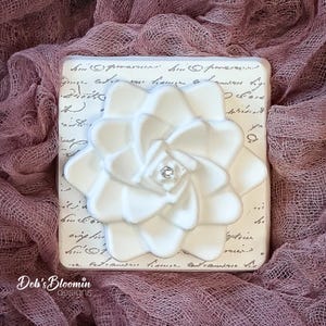 May include: A square white tile featuring a raised, sculpted white flower with a small clear crystal at its center. The tile surface is decorated with black cursive script text. The brand name Deb's Bloomin designs is printed in the bottom left corner.