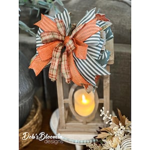 May include: A decorative wooden lantern featuring a lit LED candle inside and a large, multi-layered bow on top. The bow is crafted from ribbons in orange, grey and white stripes, and a brown plaid pattern, creating a textured, layered appearance.