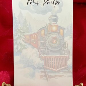 May include: A rectangular stationery card featuring a printed illustration of a steam locomotive train traveling through a snowy forest. The train is decorated with a wreath on the front. The text Mrs. Phelps is printed in black script at the top.