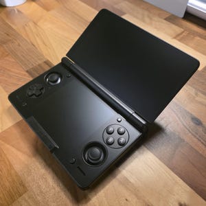 May include: A black handheld gaming console with an open top screen. The console features a directional pad, analog stick, and action buttons. The device is set against a wooden surface.