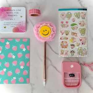 Könnte beinhalten: Eine Sammlung von Schreibwaren, darunter ein Notizblock mit Erdbeermuster, Aufkleber, ein Stift mit Smiley, Washi-Tape und ein Händedesinfektionsmittel. Die Artikel sind in Rosa-, Grün- und Weißtönen gehalten.