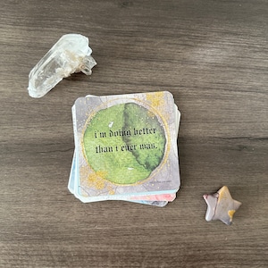 May include: A stack of oracle cards with the text "I'm doing better than I ever was." is next to a clear quartz crystal and a star-shaped stone on a wooden surface. The cards have a watercolor design with gold accents.