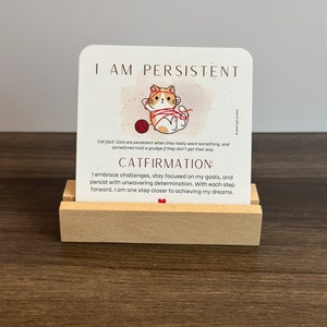 May include: A motivational card with a cartoon cat tangled in yarn. The card features the text "I am persistent" and "Catfirmation: I embrace challenges, stay focused on my goals, and persist with unwavering determination. With each step forward, I am one step closer to achieving my dreams."