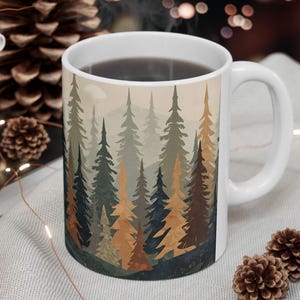 May include: White ceramic mug featuring a forest scene with evergreen trees in shades of green, brown, and blue. The mug is filled with a dark liquid and has a white handle. The mug is on a light gray surface with decorative items.