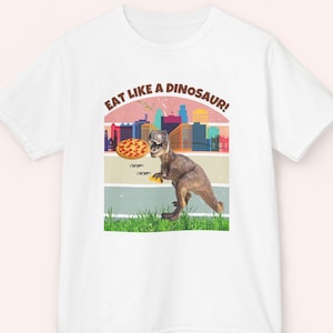 May include: White t-shirt with a graphic of a dinosaur eating pizza in front of a city skyline. The text "EAT LIKE A DINOSAUR!" is printed above the image. The dinosaur is brown and the pizza is round.