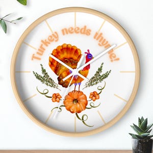 Puede incluir: Un reloj de pared redondo con marco de madera clara. La esfera del reloj presenta una ilustración de acuarela de un pavo sobre una calabaza, con el texto "Turkey needs thyme!" en naranja. Incluye ramitas y flores decorativas.