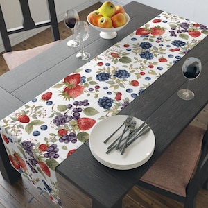 May include: A white table runner featuring a repeating botanical print of red strawberries, dark purple blackberries, and small blue blueberries with green leaves, laid across a dark wooden table with white plates and wine glasses.