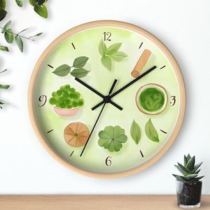 Op de afbeelding: Een ronde wandklok met een licht houten frame. De wijzerplaat is lichtgroen met aquarel illustraties van bladeren, matcha en een kaneelstokje. Zwarte wijzers geven de tijd aan. De klok staat op een houten oppervlak.