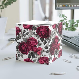 May include: A white cube-shaped notepad with a floral design featuring dark red roses and gray leaves on a white background.