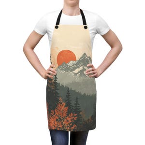 May include: Apron with a landscape design featuring a red sun, mountains, and a forest. The apron has black straps and a beige background. The bottom of the apron has a forest scene with orange and green trees.