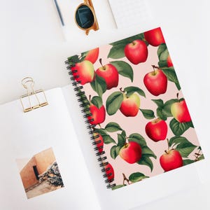 May include: A spiral bound notebook with a pink background and a repeating pattern of red apples with green leaves.