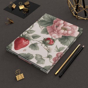 May include: A white journal with a floral design featuring pink roses, green leaves, and red strawberries. The journal is closed and lying on a dark gray surface.
