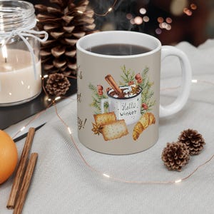 Puede incluir: Taza de cerámica blanca con asa marrón. La taza tiene un fondo beige con una ilustración de acuarela de una taza de chocolate caliente con malvaviscos, una rama de canela y un croissant. El texto "It's a Sweet Cozy Day!" está impreso en la taza. La taza está sobre una superficie blanca con una piña marrón y una cadena de luces.