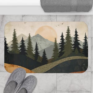 Op de afbeelding: Een rechthoekige badmat met een landschapsillustratie van donkergroene dennenbomen, grijze bergen en een grote lichtoranje zon tegen een crèmekleurige lucht. Het onderste gedeelte toont een donkergrijs vlak met subtiele witte lijntekeningen van bloemen.