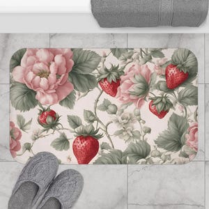 以下が含まれることがあります： イチゴとバラの模様がついたピンクとグリーンのフローラルなバスマット。マットはクリーム色の背景です。