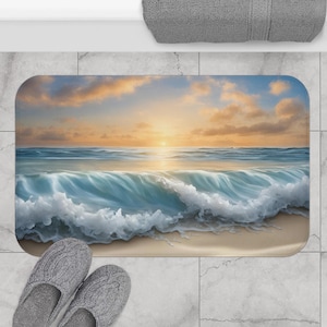 Puede incluir: Una alfombra de baño rectangular con una serena escena de playa con una puesta de sol sobre el océano. El diseño incluye olas azules, una orilla arenosa y un cielo con tonos naranja y amarillo. Unas zapatillas grises están colocadas cerca de la alfombra.