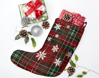 Pode incluir: Uma meia de Natal com um padrão xadrez vermelho e verde e detalhes de flocos de neve brancos. A meia está cheia de presentes embrulhados e pinhas. Presentes adicionais, pinhas e um enfeite prateado são dispostos ao redor.