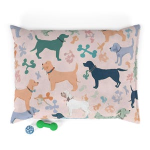 Può includere: Un letto rettangolare per cani con sfondo rosa con varie razze di cani in verde acqua, beige e blu. Forme di ossa e foglie sono sparse sul letto. Una palla blu e un giocattolo verde a forma di manubrio sono in primo piano.