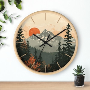 May include: Round wall clock with a light wood frame. The clock face features a mountain landscape with a setting sun, evergreen trees, and a muted color palette. Black clock hands and minimalist hour markers complete the design.