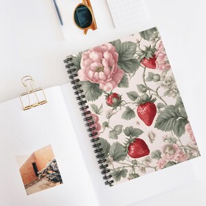 May include: A spiral bound notebook with a pink and green floral pattern featuring strawberries and roses. The background is a light cream color.