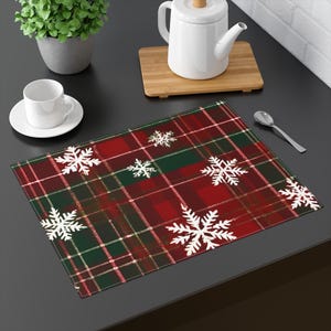 Puede incluir: Un mantel individual rectangular con un patrón de cuadros rojos, verdes y blancos. Diseños de copos de nieve blancos están dispersos por la superficie. También se ven una taza y un platillo blancos, una tetera y una cuchara.
