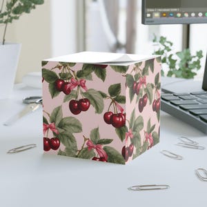 May include: A pink cube-shaped notepad with a repeating pattern of red cherries with green leaves and pink bows.