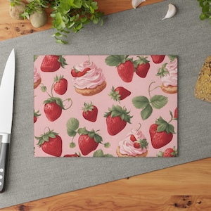 Peut inclure: Une planche à découper rose avec un motif répété de fraises et de cupcakes glacés roses. Les cupcakes sont surmontés d'une seule fraise.