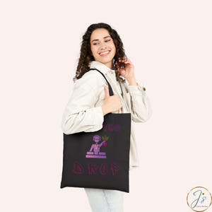 May include: A black tote bag featuring a purple graphic of a DJ wearing headphones behind a mixing console, holding a beet. The text above the graphic reads "BEET THE" and the text below the graphic reads "DROP" in a purple, stylized font.