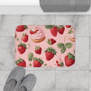以下が含まれることがあります： ピンク色の背景に赤いイチゴ、緑の葉、クリームをトッピングしたペストリーの模様が描かれた長方形のバスマット。マットはタイルの床に置かれ、灰色のスリッパと丸められたタオルが見えます。