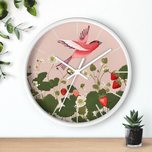 May include: A white round clock with a pink background featuring a red bird flying over a field of green strawberry plants with red strawberries and white flowers.