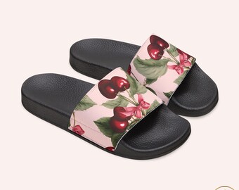 May include: A pair of black slide sandals featuring a light pink strap decorated with a repeating pattern of dark red cherries, green leaves, and small pink bows. The footbed is black with a textured surface and a molded arch for support.