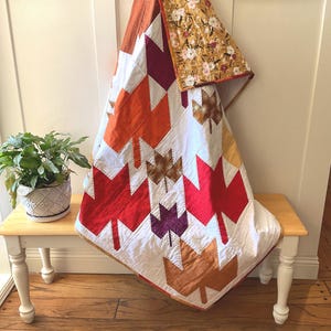 May include: A handmade quilt featuring a leaf pattern in shades of red, orange, purple, and brown on a white background. The quilt is draped over a small wooden bench, showcasing its autumnal color palette and intricate design.
