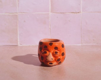 May include: Orange ceramic planter with a face and black polka dots. The planter has a closed mouth and eyes that are closed.
