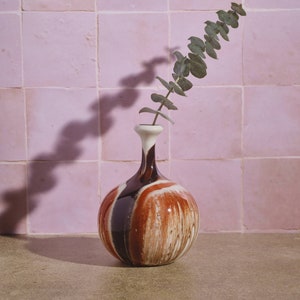 May include: A round, brown, white, and black ceramic vase with a narrow neck and a single sprig of green eucalyptus leaves.