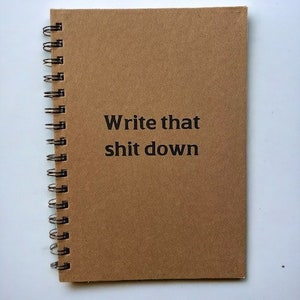 May include: A brown spiral-bound notebook with the text "Write that shit down" printed in black. The notebook has a black metal spiral binding. The paper appears to be a light brown color.