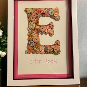 May include: A white frame with a pink mount surrounding a white background. The letter "E" is formed from colourful buttons. The text "is for Elodie" is written below the letter.