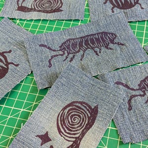 May include: Several rectangular denim fabric patches featuring dark purple ink block prints of various insects and a snail. The prints include a snail with a star, a beetle, and a long-bodied insect with multiple legs, all on blue denim.
